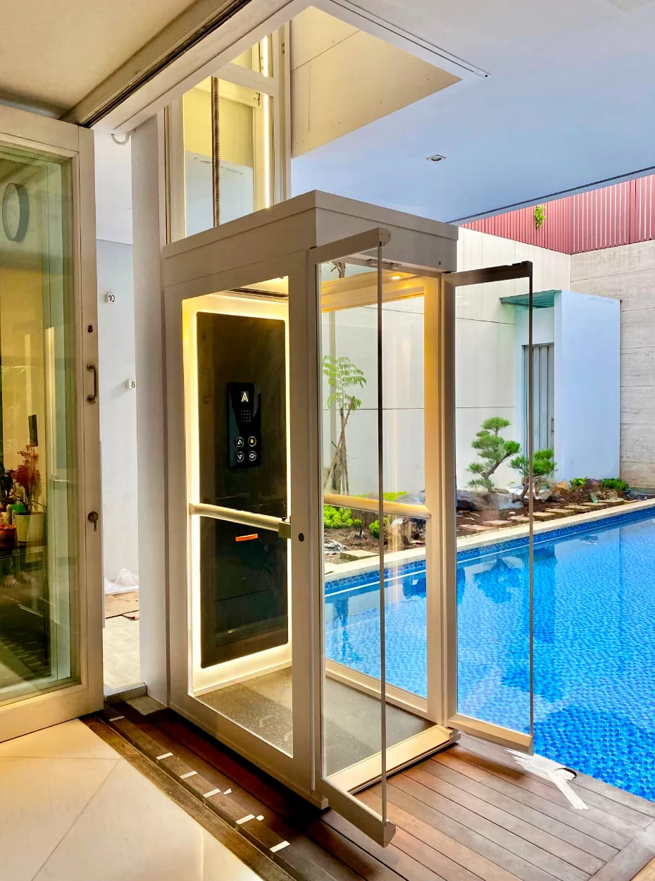 Tampak samping lift rumah Ascenda dengan shaft kaca yang terpasang di samping kolam renang dan area teras.