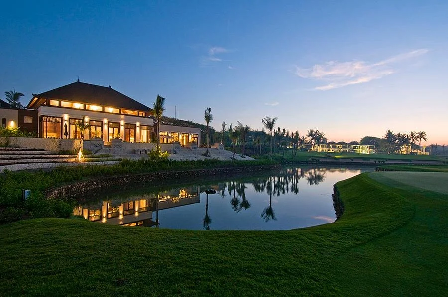 Pemandangan Bali National Golf Club Nusa Dua saat senja, clubhouse menyala dengan danau di depan lapangan.