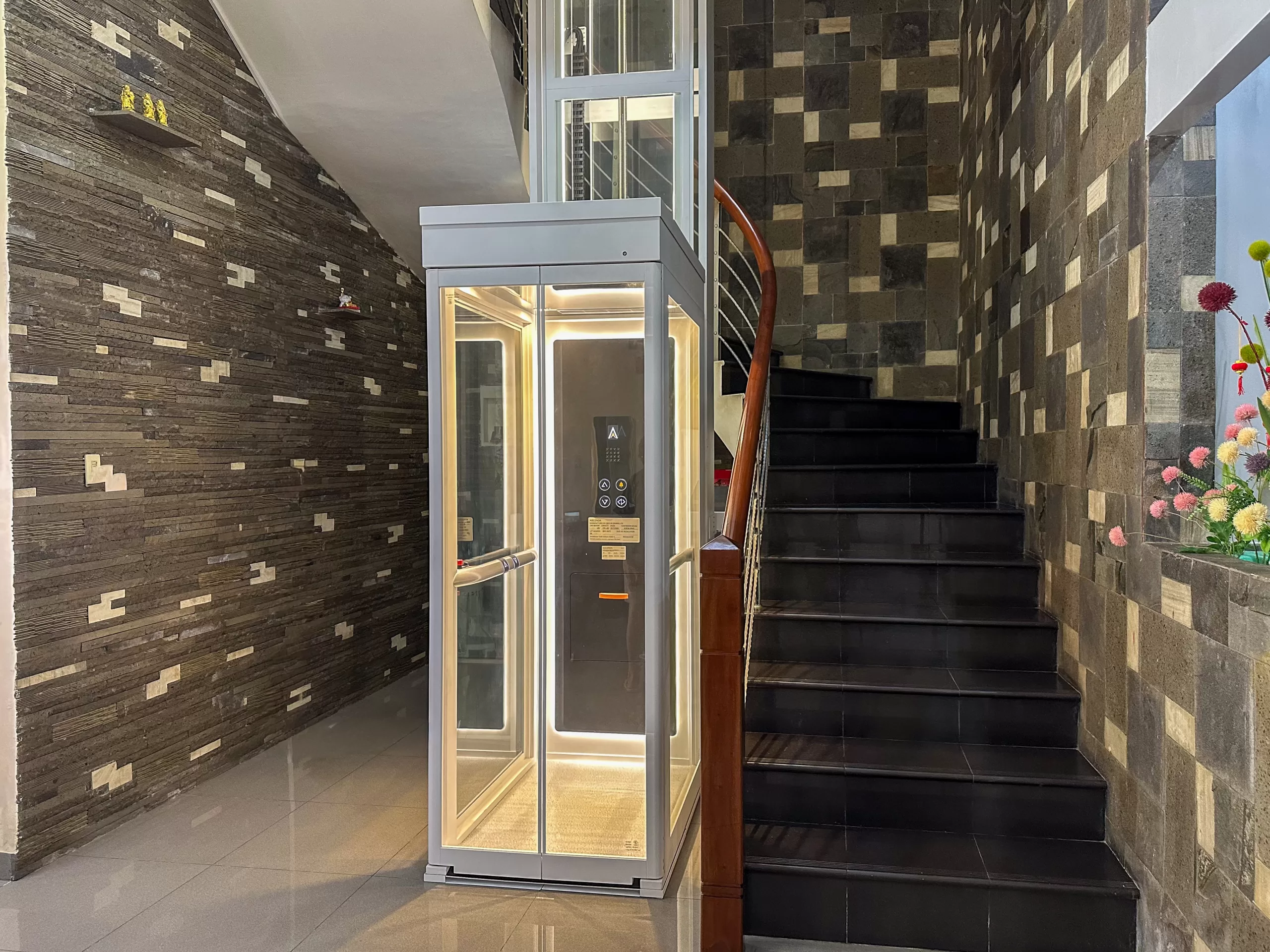 Instalasi lift rumah Ascenda Size L di Medan dengan shaft kaca dekat tangga dan dinding batu alam.