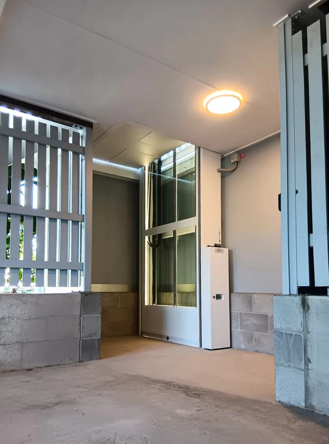 Lift rumah platform terpasang di area semi-outdoor beratap dengan dinding pelindung dan struktur bangunan di sekelilingnya.