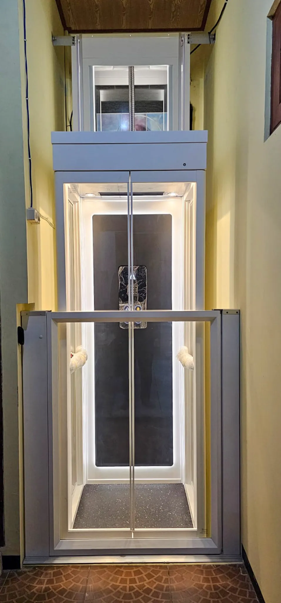 Lift rumah kecil Ascenda Size S dengan pintu kaca dan kabin berwarna putih di lorong sempit.